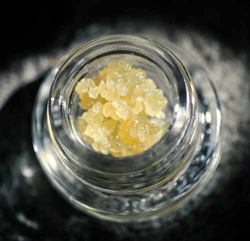 A close-up of a glass jar containing yellowish crystalline substance, set against a dark background.