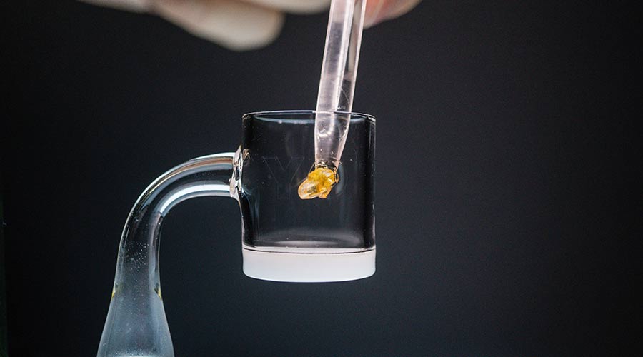 A dropper is adding a golden substance into a clear glass container, with a dark background.